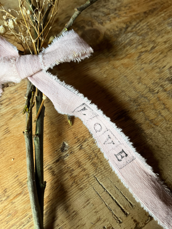 Close up of the word Love stamped onto a pink ribbon tied in the middle of a handmade twig heart decoration with dried white flowers laying on an old pine table.