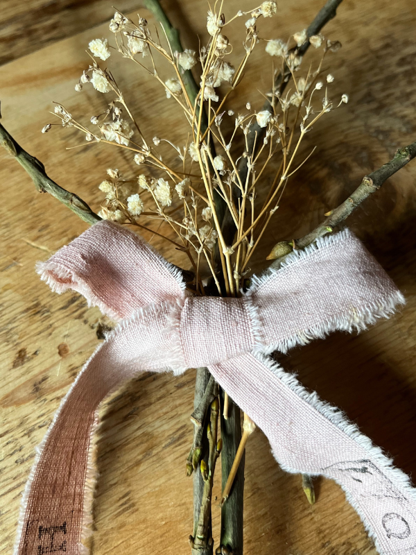 Close up of a ribbon bow tied in the middle of a handmade twig heart decoration with dried white flowers laying on an old pine table.