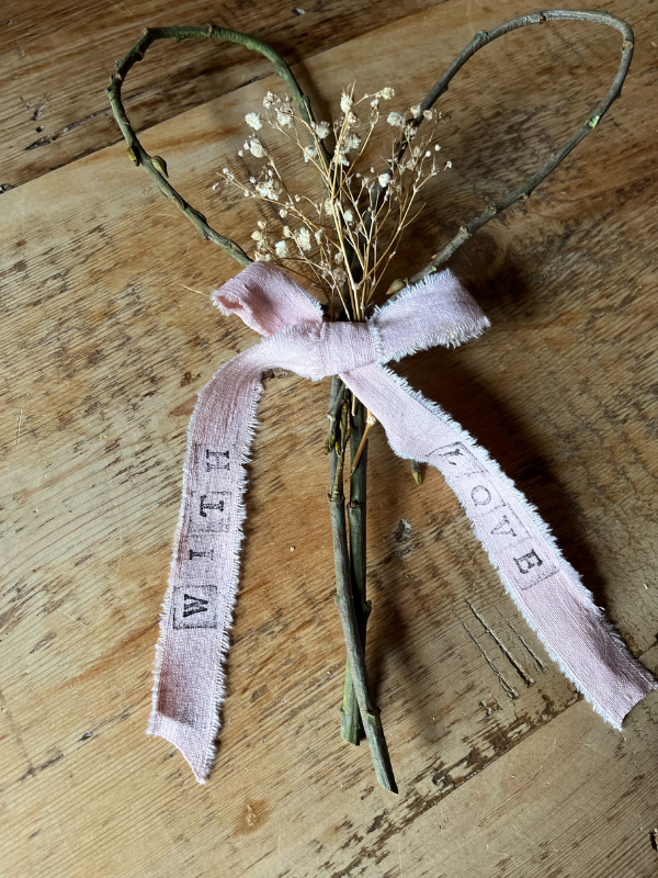 Handmade twig heart decoration with pink ribbon tied in a bow & dried white flowers laying on an old pine table. The words With Love have been stamped onto the pink ribbon.