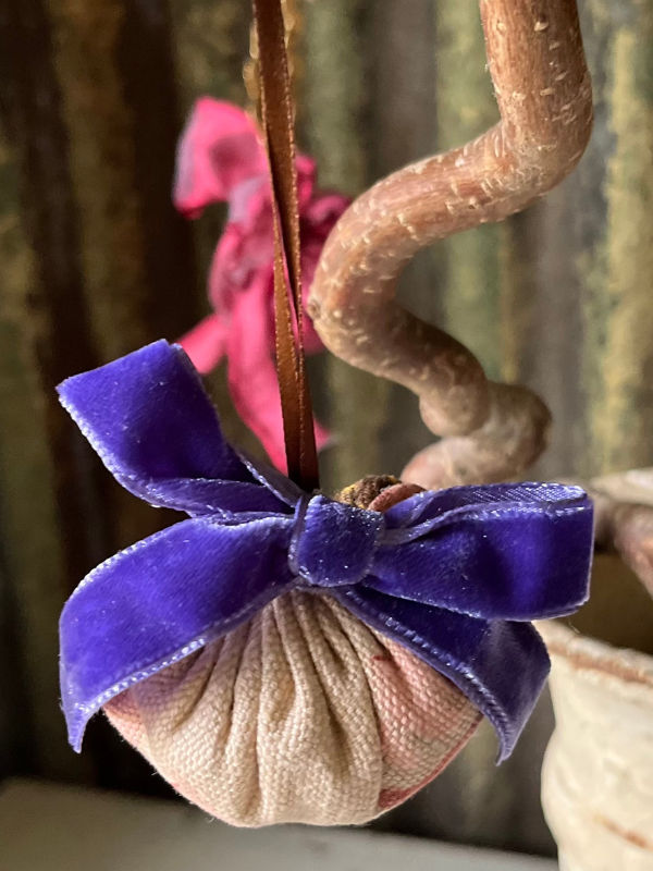 Close up of a small vintage fabric decoration hanging from a twisted tree branch. Detail of purple velvet ribbon bow on the front. Against a rusty corrugated iron background.