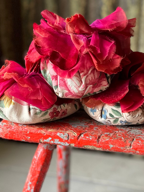 Close up of red fabric corsages on top of three handmade vintage fabric lavender filled flowers piled on top of a red wooden stool.