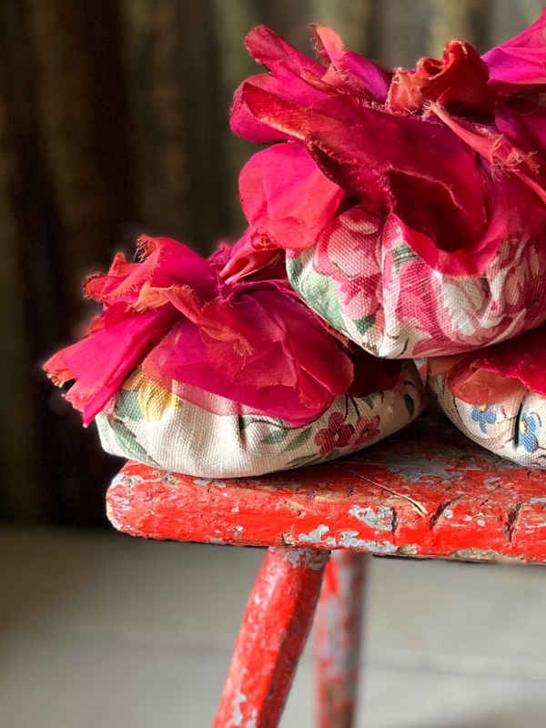 Close up of red fabric corsages on top of three handmade vintage fabric lavender filled flowers piled on top of a red wooden stool.