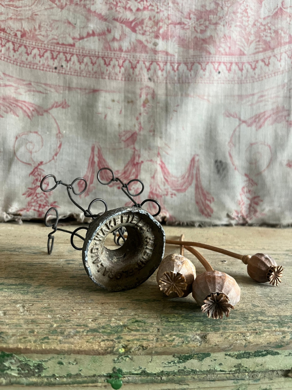 Underside of a vintage metal FloralAid flower holder against pink toile de jouy fabric with dried poppy seed heads